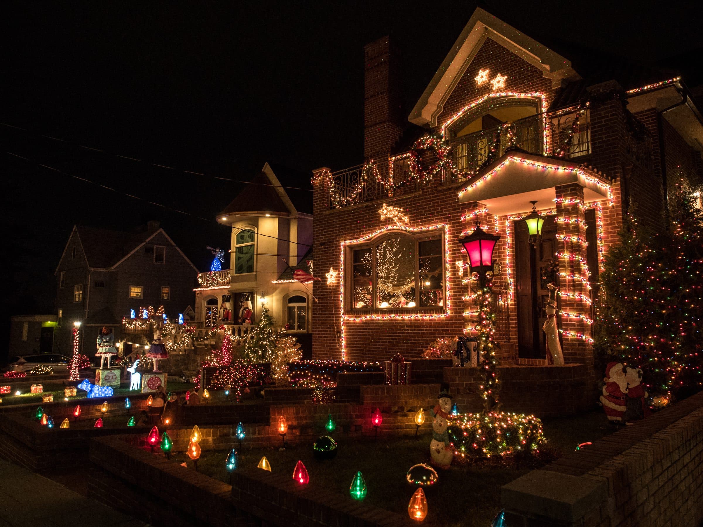 A brick house with multicolor lights on the roofline, windows, porch columns and garland, and large multicolor bulbs lining the front lawn