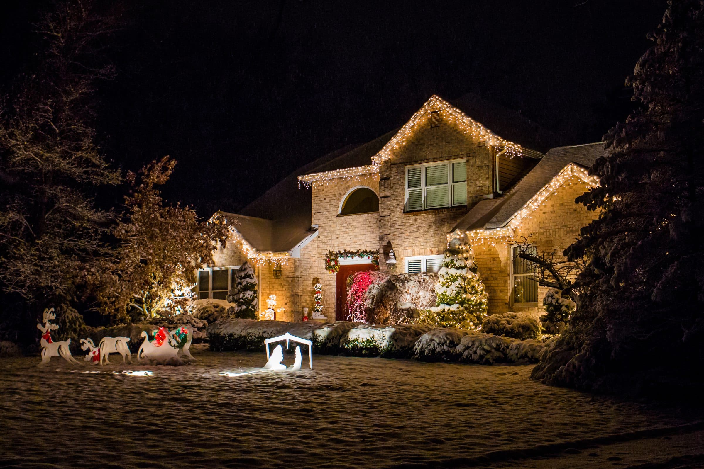 A brick house in the snow with warm white lights along the rooflines and lit trees and shrubs around the front yard