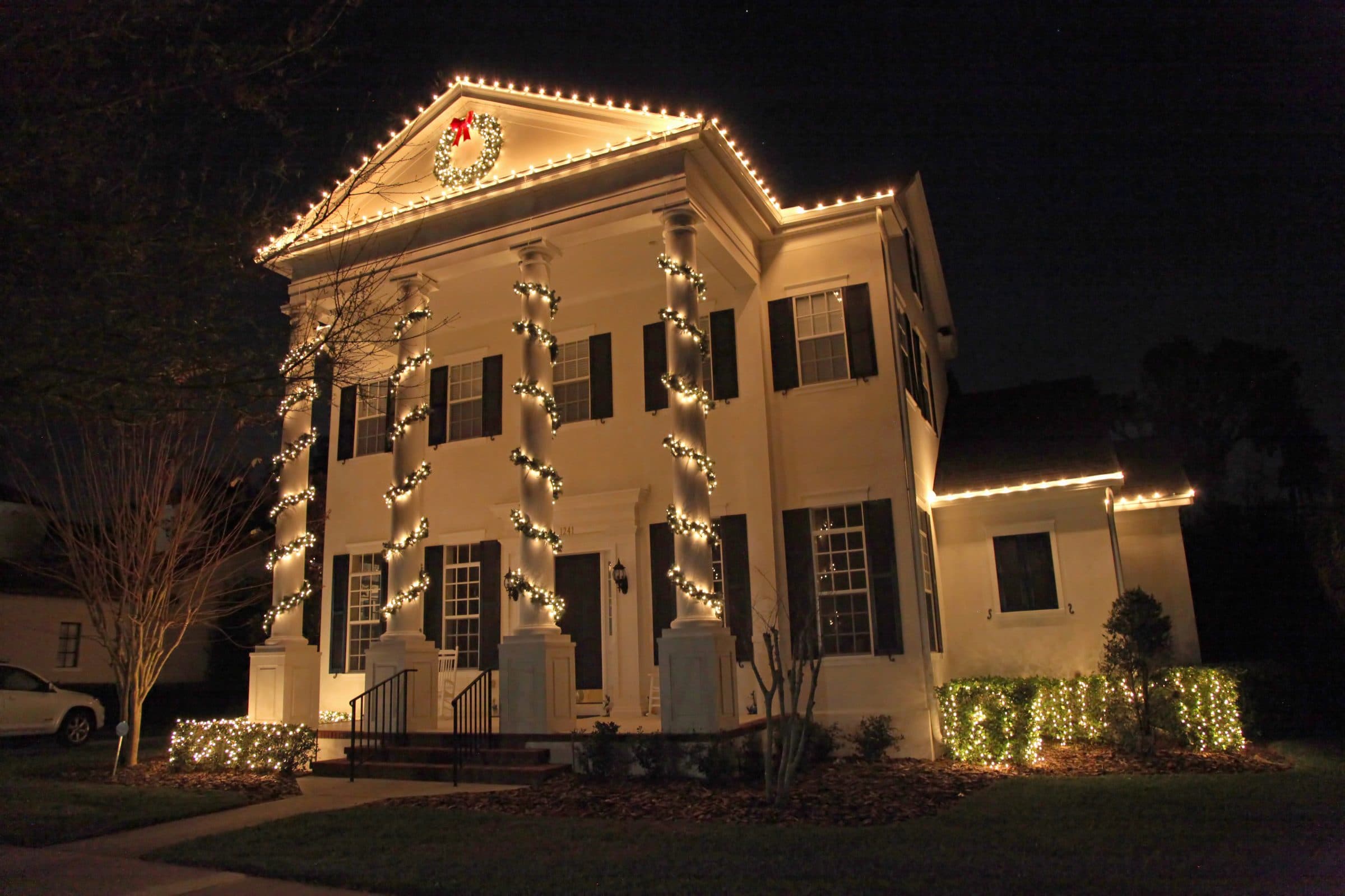 A white two-story colonial house at night with warm white lights along the roofline and pediment, lights wrapped around the porch columns and net-lit shrubs