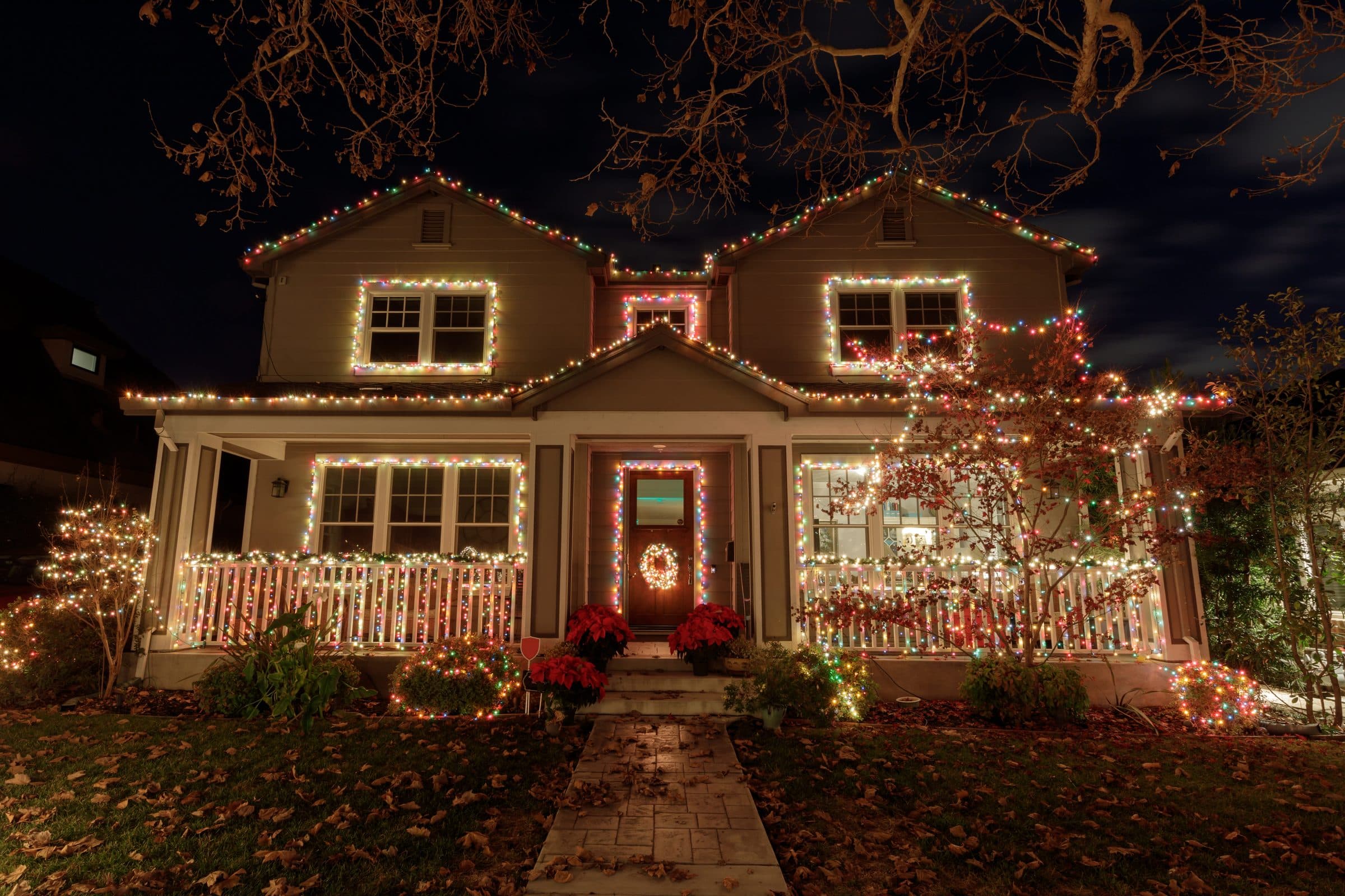 A two-story house with multicolor lights outlining the rooflines, windows, porch railing and front door, with lit bushes in the yard