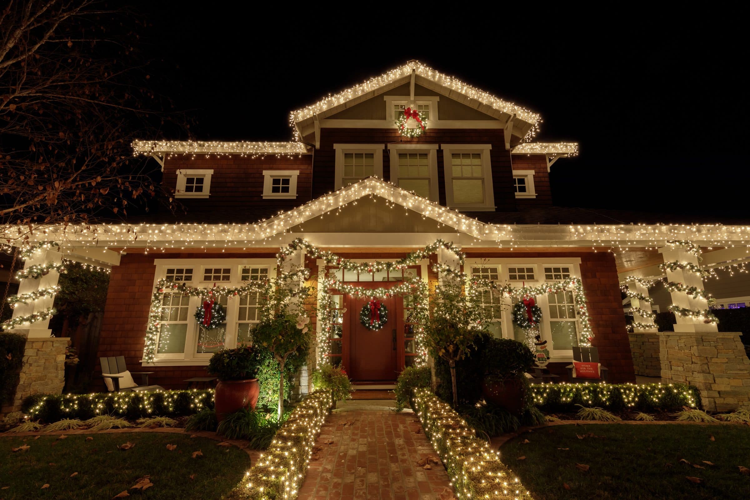 A two-story craftsman house with warm white lights along every roofline and window, wreaths, garland on the porch and lit shrubs lining the front walk