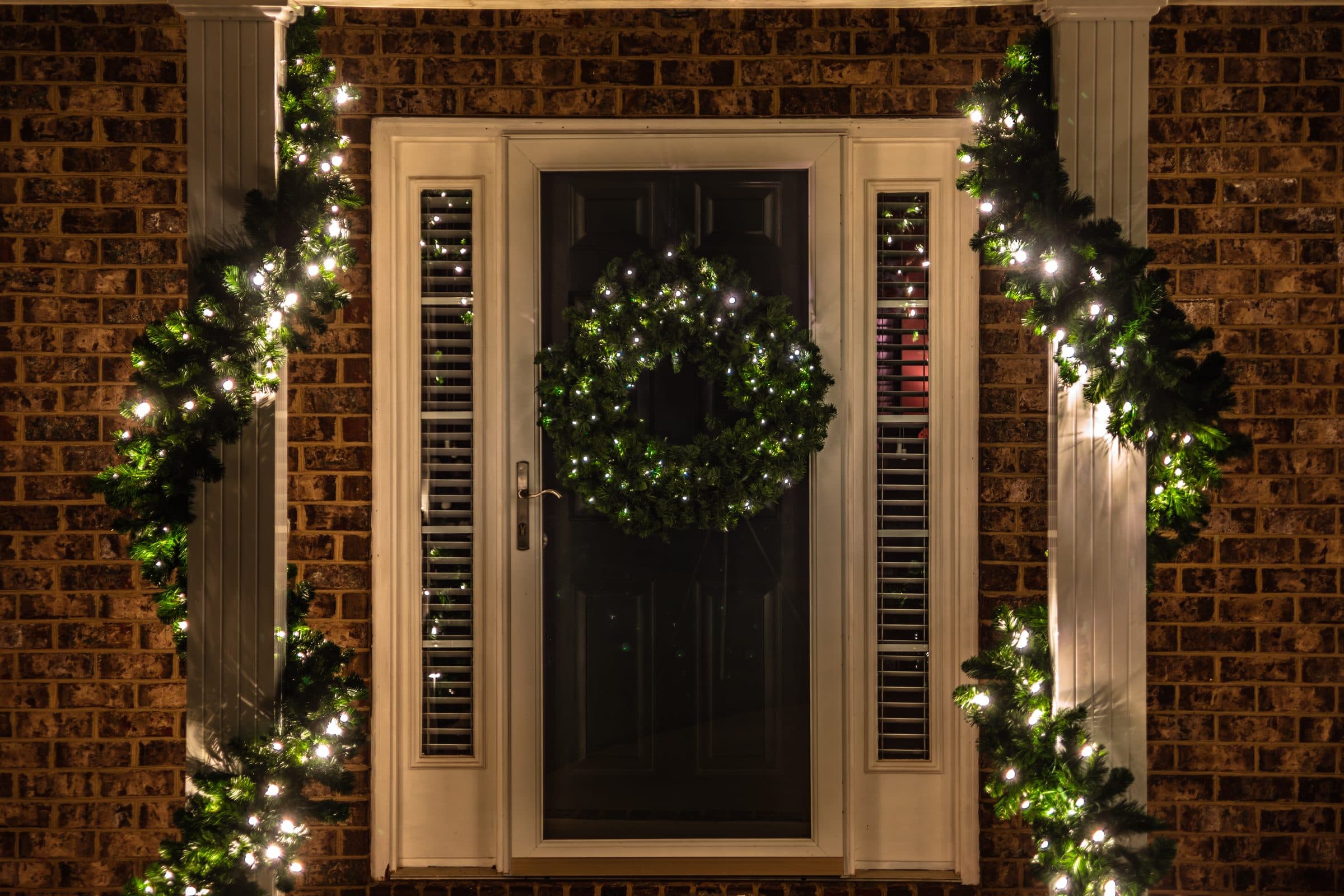 A front door with a lit green wreath, and lit garland wrapped around the white columns on either side of the door