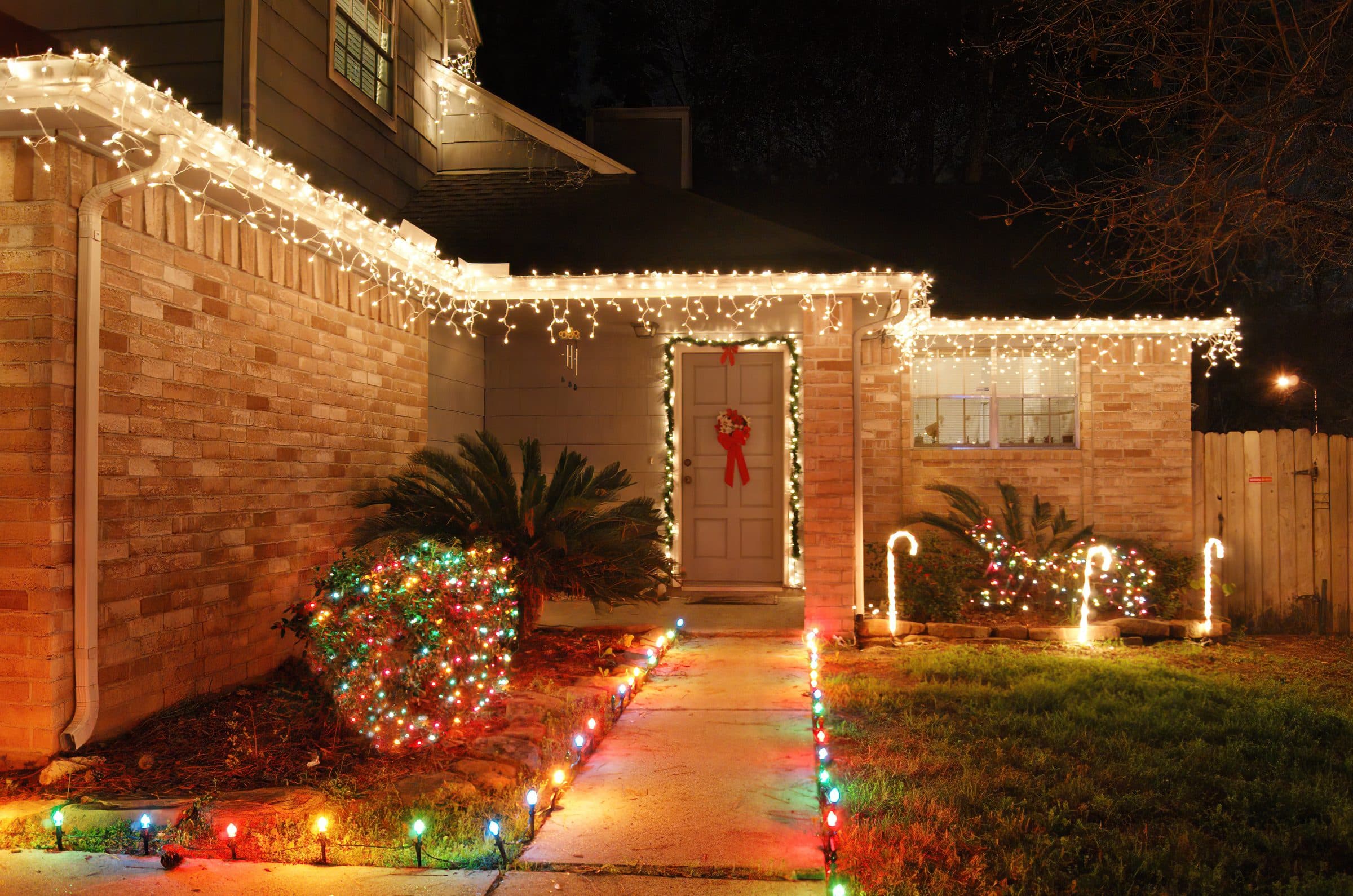A brick ranch house with white icicle lights along the roof, a multicolor-lit shrub and multicolor path lights along the front walk