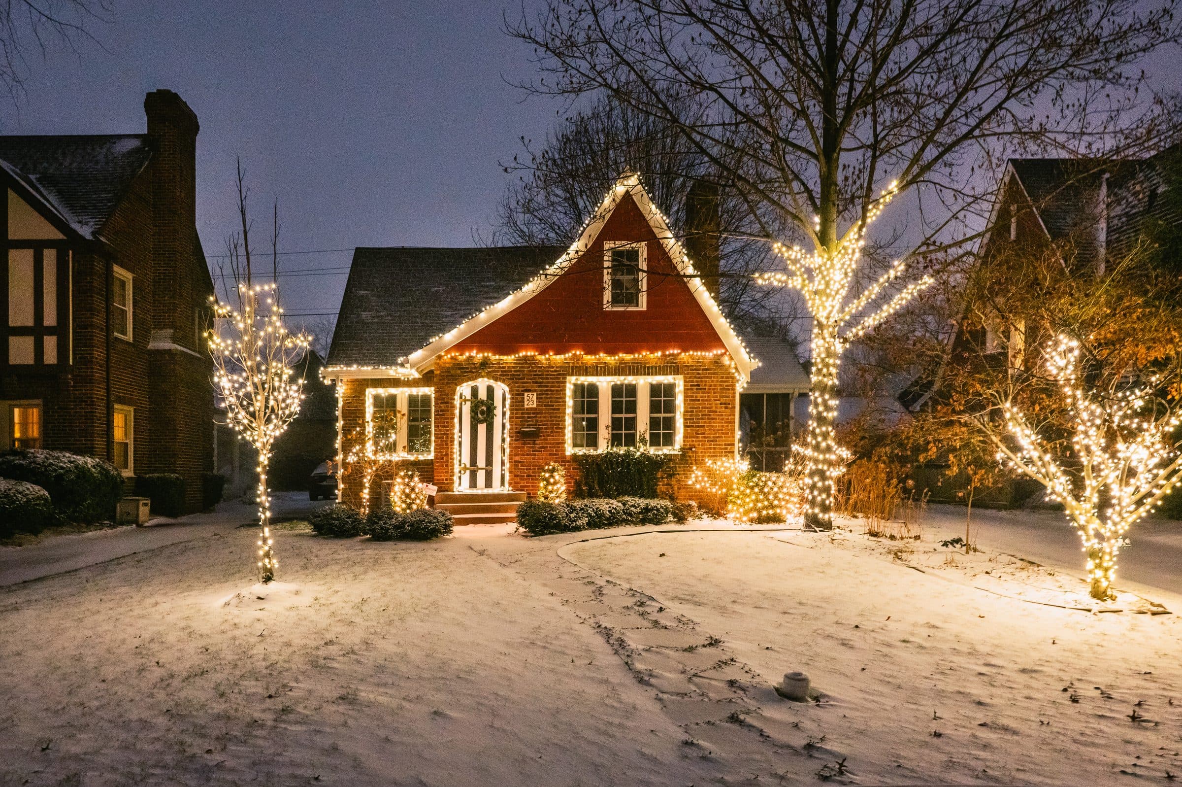 A small brick house at dusk in the snow, its roofline outlined in warm white lights, with three trees wrapped in white lights in the front yard