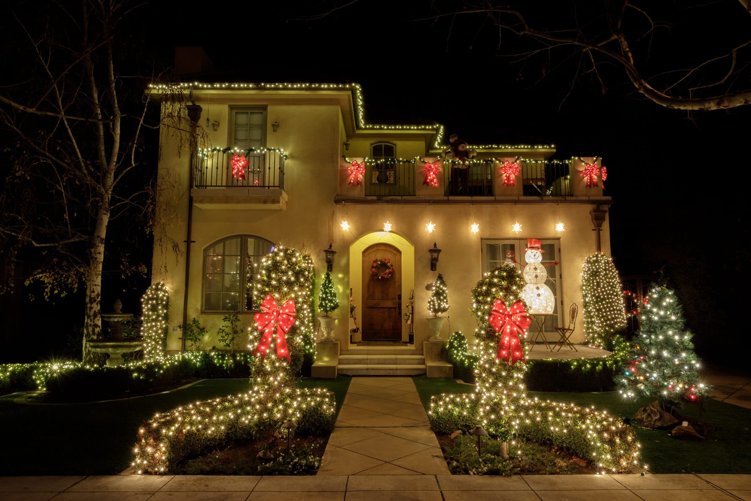 A stucco house with warm white rooflines, red bows on the balcony, and net-lit shrubs with large red bows along the front walk